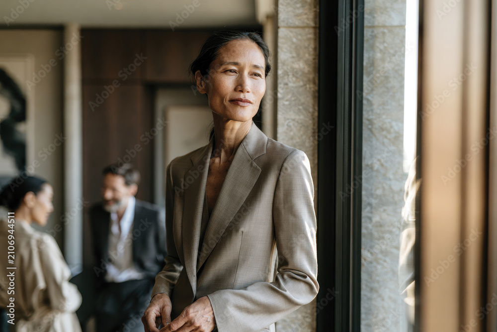 Fototapeta premium A poised woman stands by a large window, exuding confidence in a stylish suit. Her thoughtful gaze reflects determination and grace in a modern corporate environment.