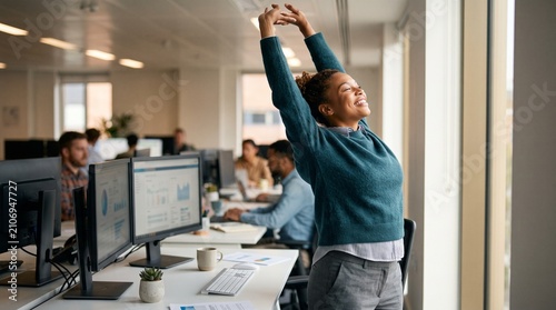 Young woman stretching arms up in joy and relief at her office desk with computers and colleagues in background
