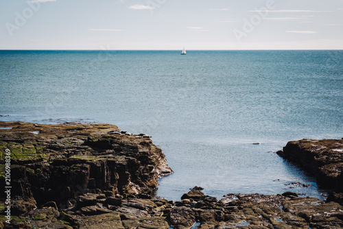 Whitley Bay UK: 13th April 2025: A sailboat glides over the water near the coastline of Whitley Bay on a clear, sunny day. The rocky shoreline frames the scene as the sea stretches toward the horizon