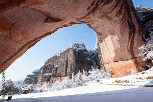 Snow-covered landscape viewed through a natural rock arch with red sandstone formations and evergreen trees.