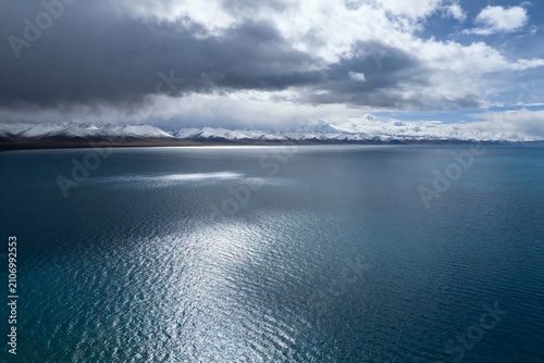 Aerial view of a vast turquoise lake with snow-capped mountains under a dramatic cloudy sky