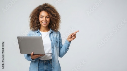 Beautiful African-American woman with laptop pointing at something on light background