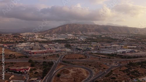 The camera shows an aerial view of a city with various buildings and roads. Hills stand in the background as the sun sets, creating warm light across the area.