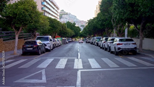 People walk on the sidewalk while cars are parked on both sides of the street. Trees line the road and buildings are seen in the background. It is early evening.