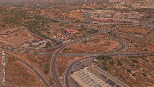Cars move along a winding road in a hilly area near buildings. The sun sets, casting an orange glow over the landscape. People can be seen enjoying the view from a distance.