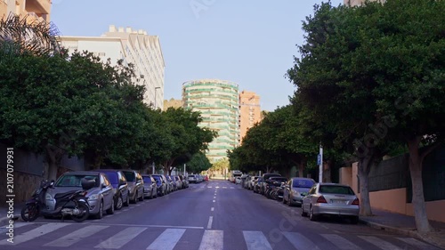 A street is shown with parked cars lined up on both sides. Trees are visible along the street. It is daytime, and buildings can be seen in the background.