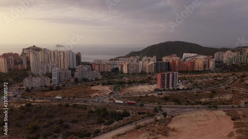 The camera shows a wide view of a city near the sea at sunset. Buildings line the area as mountains rise in the background. Clouds fill the sky, creating a mix of light and shadow.