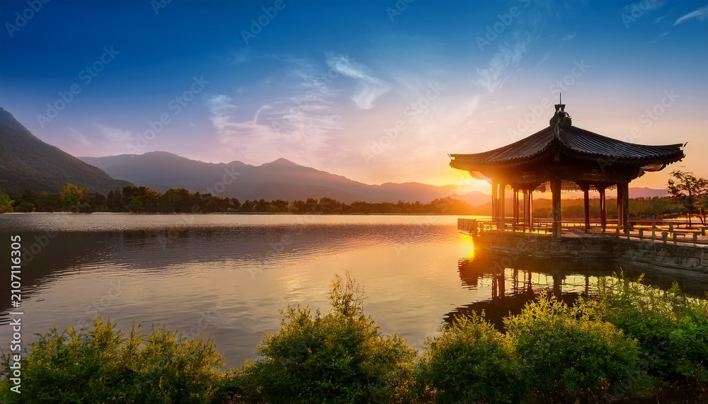 Obraz premium Serene Traditional Pavilion By A Calm Lake At Sunset With Mountains