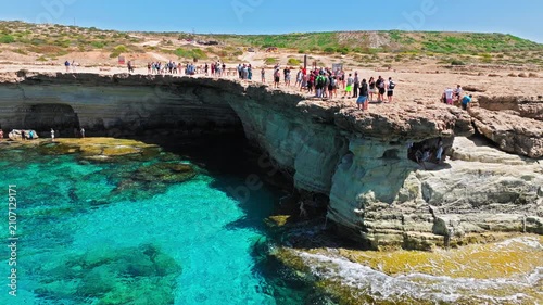 Aerial view of Cape Cavo Greco sea caves in Cyprus. Tourists explore the rocky limestone cliffs and natural cave openings above crystal-clear turquoise water and the rugged Mediterranean coastline.