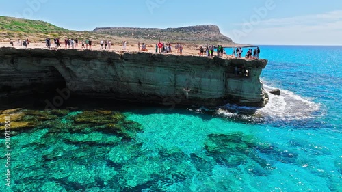 Aerial view of Cape Cavo Greco sea caves in Cyprus. Tourists visit the rocky cliff edge above crystal-clear turquoise water, natural cave openings and rugged Mediterranean coastline.