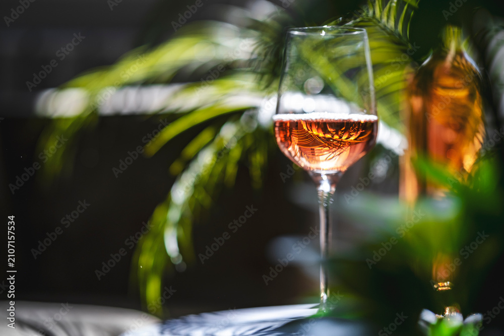 Obraz premium Pink rose wine glass in front of blurry bottle. Tropical lounge bar atmosphere and organic winemaking brand advertisement concept.