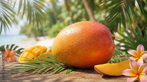 Fresh whole and cubed ripe tropical mango on a wooden table with palm leaves and frangipani flowers at a sunny beach