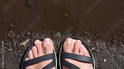 Bild auf Leinwand A top view of bare human feet in black sandals standing on muddy ground next to a brown puddle