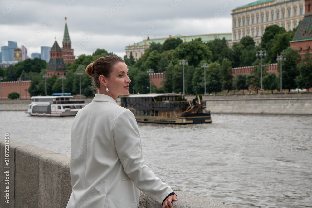 Fototapeta premium Woman glances back beside Moscow River on Sofiyskaya Embankment with Kremlin skyline in background. Urban travel, leisure, architecture, riverfront, tourism, modern lifestyle, freedom