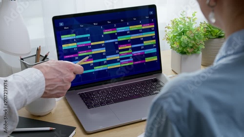 Colleague pointing pen at laptop screen displaying monthly calendar with dates for project planning. Woman reviewing schedule during time management and workplace organization.
