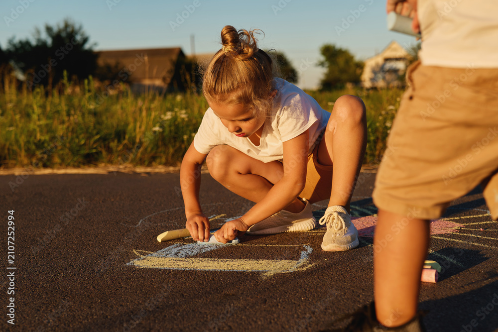 Obraz premium Two children draw pictures with chalk on the asphalt. It is evening and the sun is setting in a neighborhood with green fields