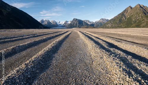 Wide gravel path leading through a vast flat valley toward distant snow-capped mountains under a clear blue sky in a remote wilderness area