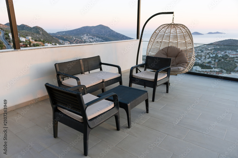 Obraz premium Modern outdoor patio seating and a hanging rattan chair on a tiled terrace overlooking a coastal town and sea at sunset. Comfortable cushions, panoramic mountain and ocean views create a relaxing mood