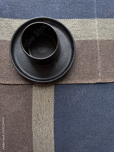 Minimal black ceramic cup on striped linen textile. Handmade stoneware and natural fabric create a simple composition with neutral tones for home decor, dining, interior and lifestyle projects.