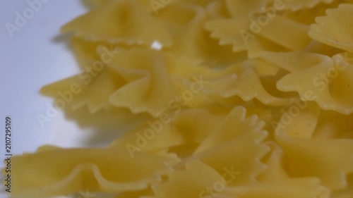 Close-up of raw farfalle bow-tie pasta rotating on a white plate, showcasing its textured shape and golden yellow color under bright studio lighting.
