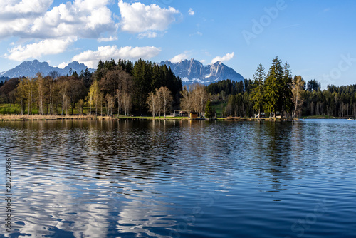 spring Tyrolean lake Schwarzsee with reflection of blue sky and white clouds, in the background the Wilder Kaiser Alpine massif with remnants of snow
