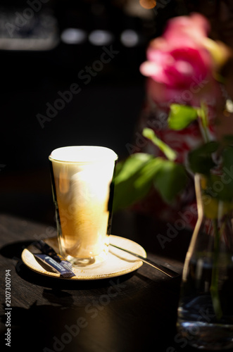 Freshly prepared hot cappuccino served in a clear glass with creamy milk foam on a ceramic saucer. Natural sunlight and soft floral bokeh create a warm outdoor café atmosphere.