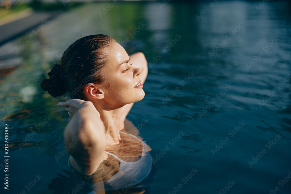 Fototapeta premium Young woman with brunette hair relaxes in an outdoor swimming pool with her eyes closed and hands behind her head enjoying a peaceful moment on a sunny day.