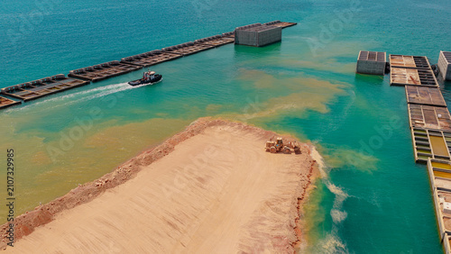 Elevated view showing a tugboat moving past a developing harbor area with large concrete structures in the water and a wheel loader working on a reclaimed landmass