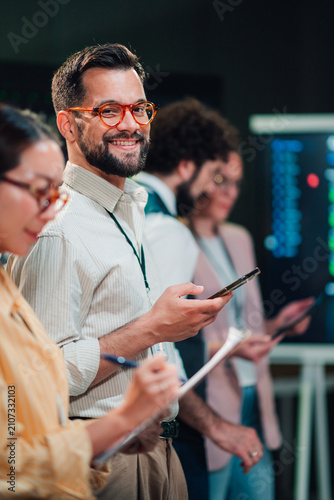 Smiling businessman in orange glasses standing with diverse team, holding smartphone and participating in a conference meeting