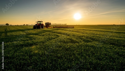 Red tractor pulling agricultural equipment through a sprawling green field under a bright golden sunset sky landscape