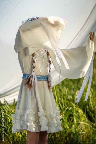Foto A young girl wearing a detailed white traditional folk dress with blue embroidery and long wide sleeves poses in a grassy field outdoors while holding a translucent fabric banner