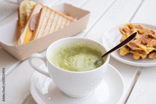 Matcha latte with milk and foam in a white mug on the table, waffles and a sandwich.