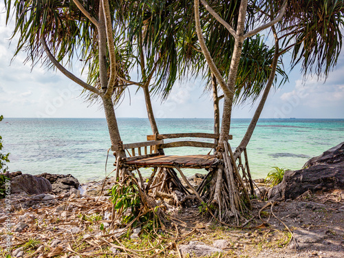 Rustic wooden bench offering a shaded resting spot beneath pandal trees with sprawling roots. Overlooking the clear turquoise ocean and island horizon on a tropical beach in Karimunjawa. Jawa Tengah