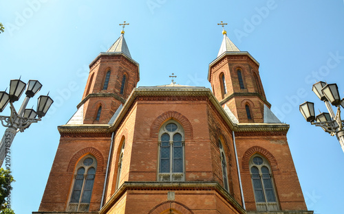 Low angle, monumental view of the historic red brick Armenian Church in Chernivtsi, Ukraine, highlighting its Gothic-revival towers, twin spires, and arched windows under a blue summer sky