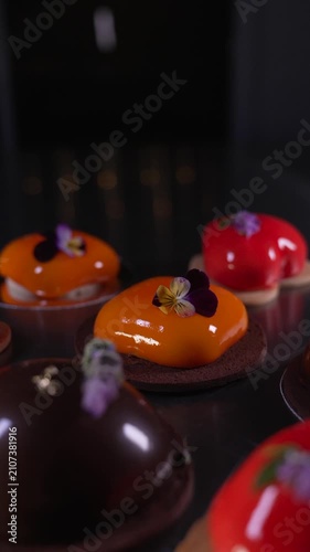 Close up of beautifully crafted assorted mousse pastries in chocolate, red, and orange with shiny mirror glazes and elegant decorations displayed on a table, haute cuisine confectionery