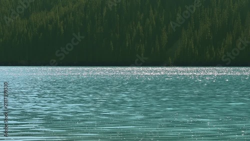 View across a turquoise colored mountain lake with the summer sky flashing on small waves. Distance evergreen trees are side lit by the sun.
