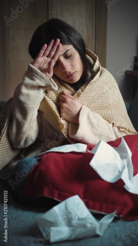 Young Hispanic woman sitting on her sofa at home with fever and chills. Health lifestyle video representing flu symptoms, illness, recovery, self-care and resting at home.
