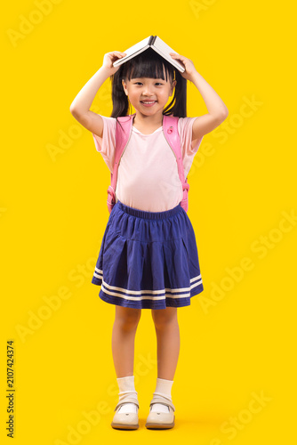 Fotografie A cute Asian female student stands against a yellow background with a book on her head, happily and cheerfully welcoming the new semester