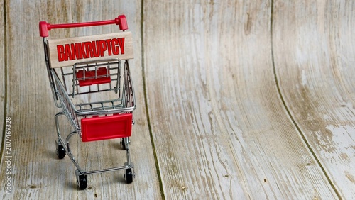 Photography An empty shopping cart with a wooden block reading BANKRUPTCY on a textured background