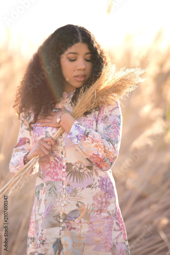 A young dark-skinned woman stands among the golden reeds. Her floral-print dress shimmers in the warm sunlight. Her curly hair flows gently over her shoulders.