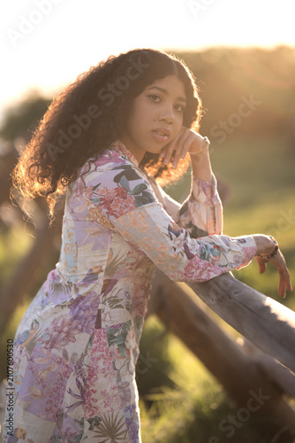 A young dark-skinned woman stands among the golden reeds. Her floral-print dress shimmers in the warm sunlight. Her curly hair flows gently over her shoulders.