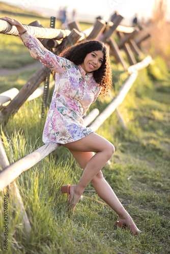 A young dark-skinned woman stands among the golden reeds. Her floral-print dress shimmers in the warm sunlight. Her curly hair flows gently over her shoulders.