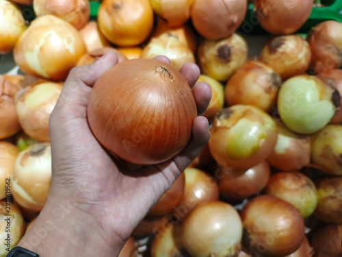 Close-up of a hand holding a fresh brown onion with a pile of onions in the background at a traditional market. Concept of selecting fresh produce, grocery shopping, agriculture, healthy ingredients
