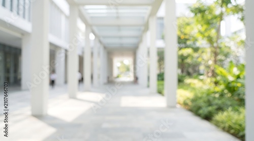 Blurred outdoor corridor walkway in modern office building with white pillars and lush green garden view for architectural background design and commercial real estate presentations
