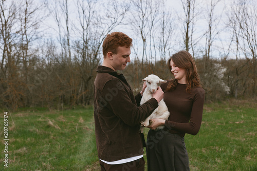 Fotografie A couple outdoors in a field interact with a goat kid, showing affection, smiles