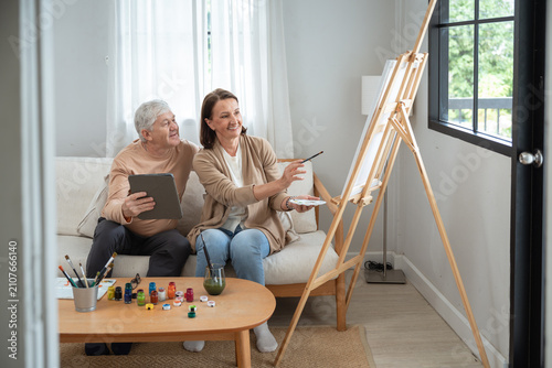 Senior Couple Sharing a Painting Hobby at Home as a Retired Woman Works at an Easel Beside Her Partner Using a Tablet During Their Active Retirement