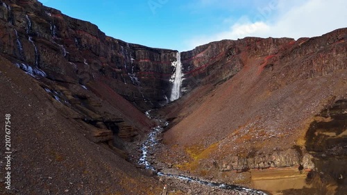 Wide aerial view flying into Hengifoss canyon with red basalt cliffs in Iceland on a partly sunny day