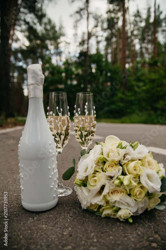 A bottle of champagne decorated for a wedding stands on the asphalt in the middle of the forest next to two crystal glasses and a bouquet of white roses
