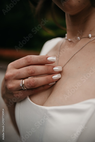 A bride in a white dress holds a piece of jewelry hanging around her neck with her hand, which has a beautiful white manicure on her fingers