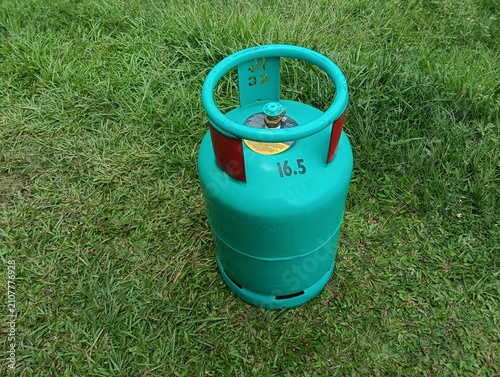 A green 16.5 kg LPG gas cylinder placed upright on a grassy field outdoors. The steel liquefied petroleum gas cylinder features a protective ring around the valve . in malaysia